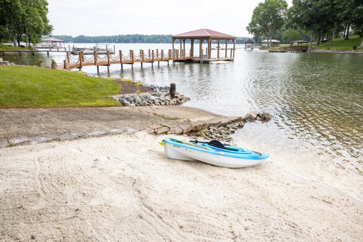 Lake-Norman-vacation-retreat-Kayak | Southern Charm Retreats A blue and white kayak rests on a sandy beach next to a calm lake with a wooden boat dock and covered pavilion.