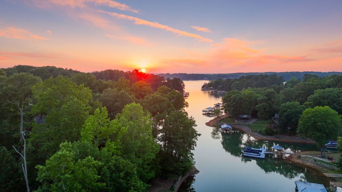 DJI_20260611203157_0664_D | Southern Charm Retreats Sunset over a lake surrounded by dense green trees, with houses and docks along the shoreline.
