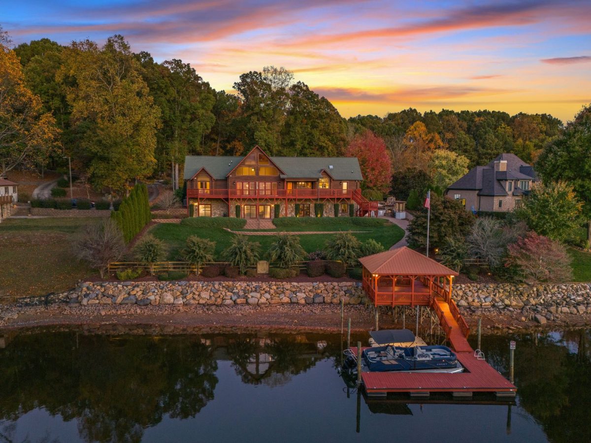 1-web-or-mls-norcross twilight-3 | Southern Charm Retreats Lakeside wooden house with red balcony railings on a grassy yard, autumn trees, and a rocky shoreline by calm water at sunset.