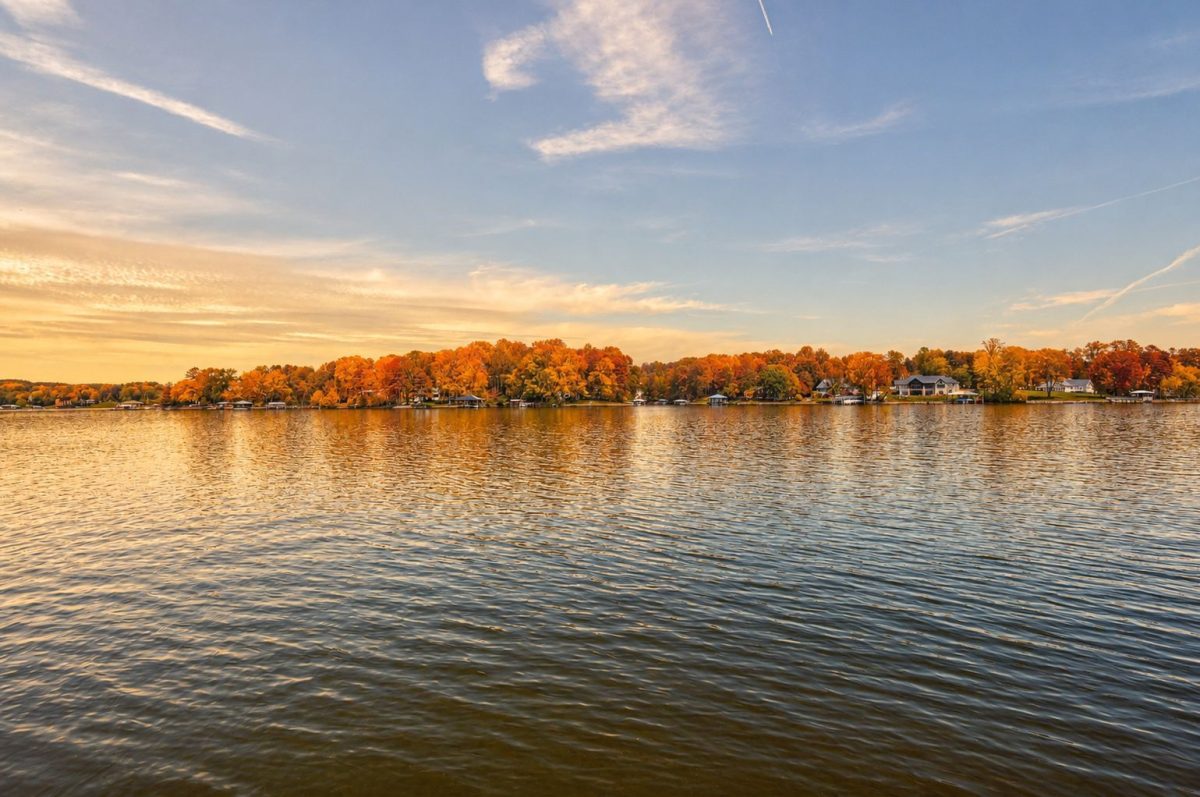 LKN fall | Southern Charm Retreats Calm lake at sunset with a tree-lined shoreline in brilliant autumn colors and houses along the water.